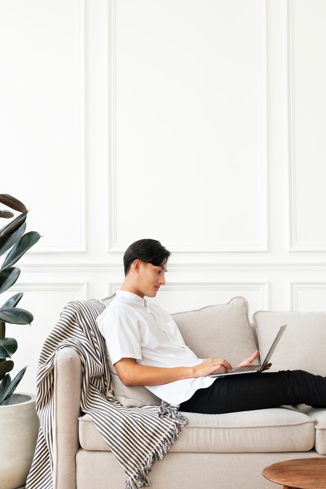 Man Using A Laptop On The Sofa, throw blanket on his back 