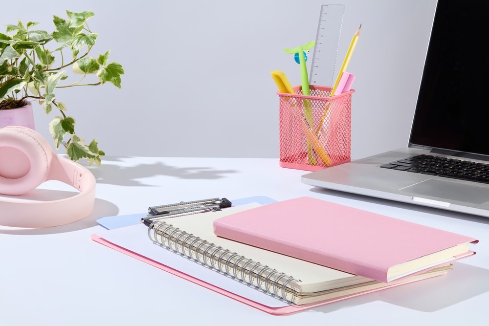  In the middle of white tabletop contains some notebook and clipboard, next to a laptop, pen pot, and a pink headphone. 