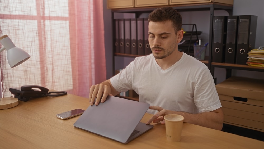 a young man working on a laptop in a modern office with pink curtains and organized shelves in the background.