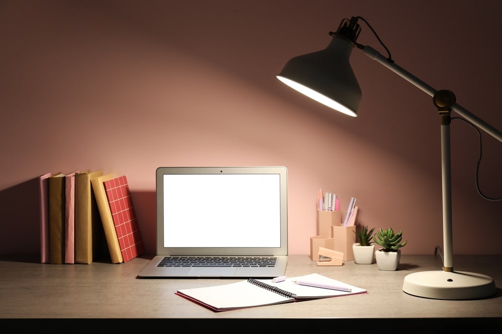 Workplace with modern laptop, notebook and glowing lamp near pink wall