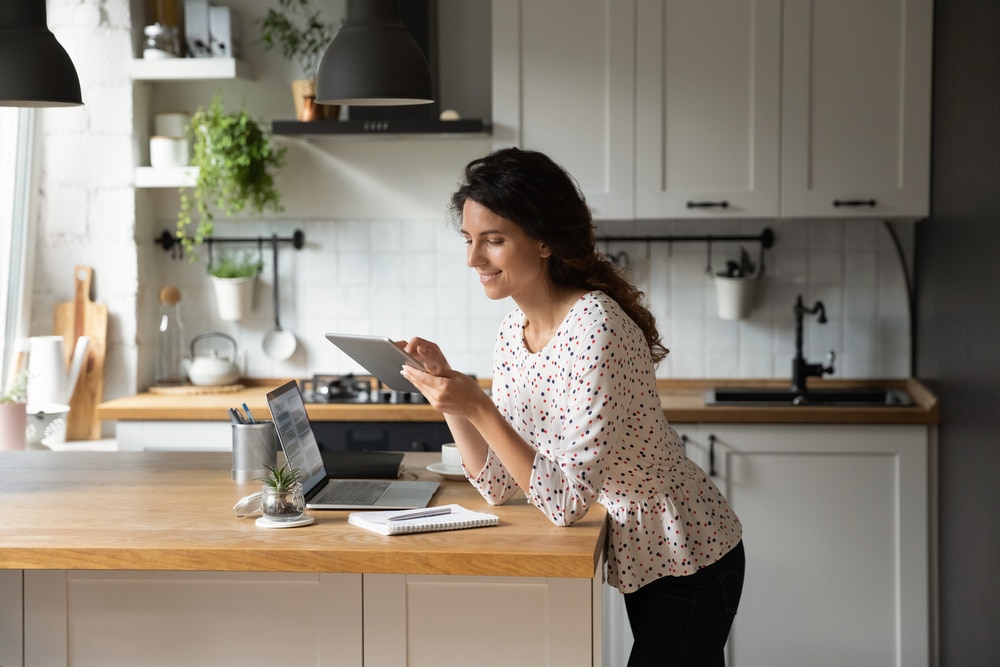 a woman working on iPad and laptop, standing in kitchen 